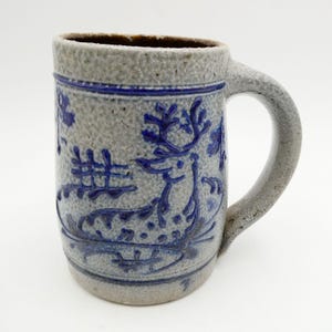 May include: A gray stoneware mug with a textured, speckled surface and a large handle. The exterior features a cobalt blue embossed design of a deer with antlers, surrounded by stylized foliage and geometric patterns encircling the rim.