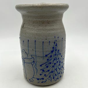 May include: Gray stoneware jar with blue hand-painted Christmas tree and figure design. The jar has a cylindrical shape with a slightly flared rim. The text "BEAVER FALLS" is stamped on the side. A charming, rustic holiday decoration.