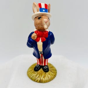 Può includere: Una statuina in ceramica di un coniglio vestito con abiti patriottici. Il coniglio indossa un cappotto blu, un papillon rosso e un cappello a cilindro con strisce rosse, bianche e blu e una stella. Il coniglio è su una piccola base rotonda erbosa.