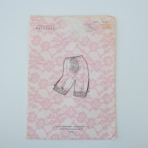May include: A vintage pattern for a pair of white lace panty girdle. The pattern is printed on a pink background with a lace pattern. The pattern number is 6926 and the price is $1.00. The pattern is from Daleres of St. Paul, Original.