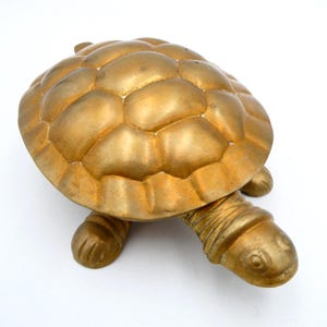 May include: A gold-coloured turtle figurine with a textured shell and detailed legs and head. The turtle is made of a solid material and sits on a white surface. The figurine is a decorative item.