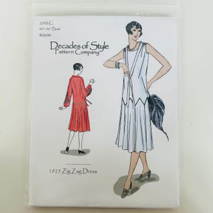 Peut inclure: Une pochette de patron de couture pour une robe 1925 Zig Zag Dress de Decades of Style Pattern Company. La couverture présente des illustrations d'une personne portant une robe blanche sans manches et une robe rouge à manches longues avec des détails d'ourlet en zigzag.