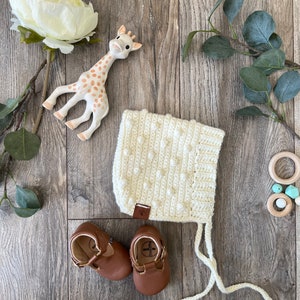 Può includere: Un berretto per neonato in crochet bianco con pompon bianchi, un giocattolo da dentizione a forma di giraffa e due scarpine per neonato in pelle marrone.