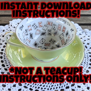 May include: A white teacup with a green saucer, decorated with playing cards. The teacup is upside down, and the text "The Cup of" is visible on the side. The text "*Not a teacup! Instructions only!" is written in red and black letters on a white background.