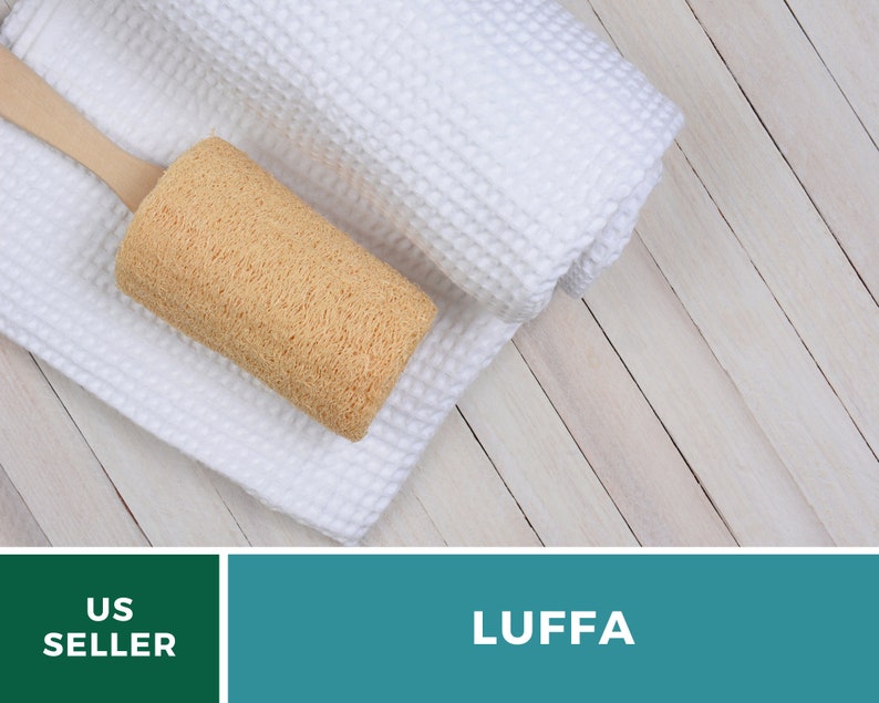 Puede incluir: Una esponja de luffa natural con un mango de madera, descansando sobre una toalla blanca. La luffa es de color marr&oacute;n claro y tiene una superficie texturizada. La toalla est&aacute; enrollada y tiene un tejido de gofre. El fondo es una superficie de madera clara.