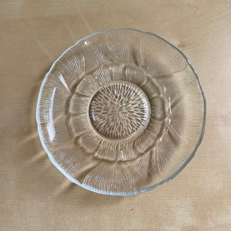 Pu&ograve; includere: Un piattino di vetro trasparente con un design in rilievo testurizzato che ricorda un fiore. Il piattino ha un pozzetto centrale per una tazza.