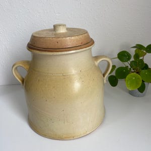 Puede incluir: Un tarro de cerámica beige con tapa. El tarro tiene dos asas y una forma redondeada. La tapa es de un tono beige más claro y encaja perfectamente. El tarro probablemente se utiliza para el almacenamiento y tiene un aspecto rústico y hecho a mano.