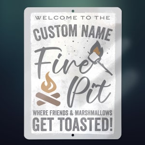 May include: White metal sign with the text "Welcome to the [CUSTOM NAME] Fire Pit" and a graphic of a campfire and a marshmallow on a stick. The text "Where friends & marshmallows get toasted!" is below the main text.