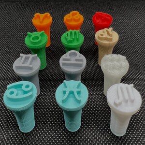 Könnte beinhalten: Zwölf bunte Stempel mit verschiedenen Sternzeichen-Symbolen und Farben. Die Stempel sind aus Kunststoff und haben eine runde Oberseite mit einem erhabenen Symbol. Die Unterseite der Stempel ist konisch geformt.