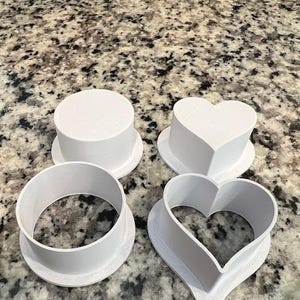May include: Four white plastic cookie cutters: a circle, a heart, a smaller heart, and a larger circle.