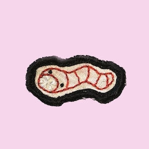May include: Embroidered patch featuring a stylized worm design. The worm is outlined in red thread, with a cream-colored body and a black border. The worm's face has a cluster of white beads. The patch is on a pink background.