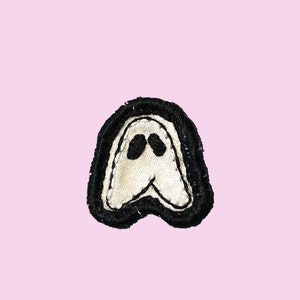 May include: A small, embroidered patch featuring a ghost design. The ghost is white with black eyes and a black outline, set against a black border. The patch is likely for clothing or accessories.