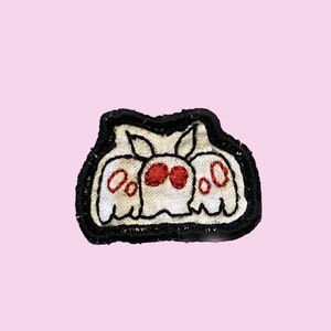 May include: A fabric patch featuring an embroidered moth design. The moth is white with prominent red eyes and red circular markings on its wings. The entire design is outlined in thick black thread, creating a bold contrast against the white fabric.