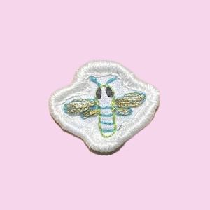 May include: A small embroidered patch featuring a stylized bee with metallic gold wings, a light blue and lime green striped body, and black eyes, all set against a white fabric background with a thick, white, scalloped embroidered border.