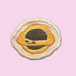 May include: Embroidered patch featuring a smiling black hole design. The patch has a white border and a black center with orange and yellow details. The patch is on a light pink background.