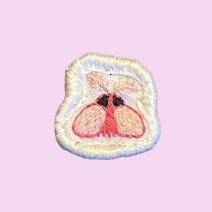 May include: A small, square-shaped embroidered patch featuring a stylized moth design. The moth has vibrant pink wings, two prominent black eyes, and light-colored antennae. The entire patch is bordered by a thick, white, raised stitched edge.