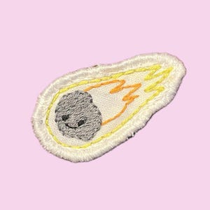 May include: Embroidered patch featuring a gray asteroid with a smiling face, trailing yellow and orange flames. The patch has a white border with yellow stitching, set against a light pink background. The design is whimsical and playful.