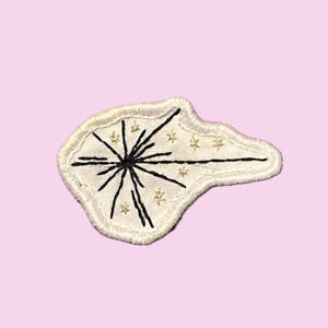 Pode incluir: Patch bordado com fundo branco e design de explosão estelar preta. O patch tem uma borda creme e pequenos detalhes de estrelas douradas. O patch está em um fundo rosa claro.