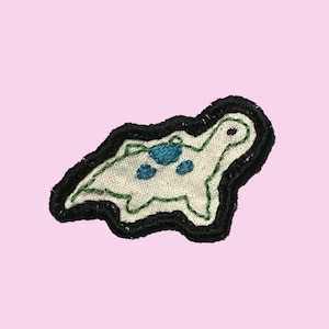 May include: An embroidered patch shaped like a turtle with an off-white fabric base, a thick black stitched border, green thread outlining the body, and three small blue embroidered spots on the shell area, featuring a simple stitched eye.