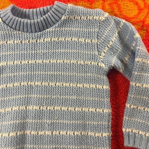 Può includere: Un maglione lavorato a maglia blu chiaro con strisce bianche orizzontali. Il maglione ha un girocollo e maniche lunghe. Il motivo a maglia è visibile, creando un aspetto strutturato. Lo sfondo è arancione e rosso vibrante.