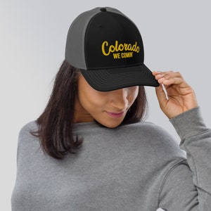 Puede incluir: Una gorra de camionero negra y gris con un logotipo bordado en oro que dice "Colorado We Comin'"