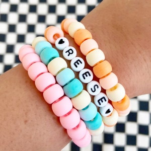 May include: A set of four colorful beaded bracelets with the name "REESE" spelled out on one of the bracelets. The bracelets are made of pastel pink, yellow, blue, and orange beads.