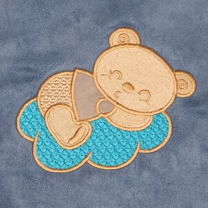 Può includere: Un orsacchiotto di peluche ricamato marrone con un occhio chiuso e un sorriso è sdraiato su una nuvola ricamata blu e marrone. La nuvola ha un centro turchese con un motivo di piccoli cuori.