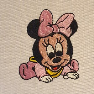 Può includere: Immagine ricamata di Baby Minnie Mouse. Il personaggio dei cartoni animati è in posizione di gattonamento, indossa un completo rosa e un bavaglino giallo. Minnie ha un fiocco rosa sulla testa e un'espressione sorridente.