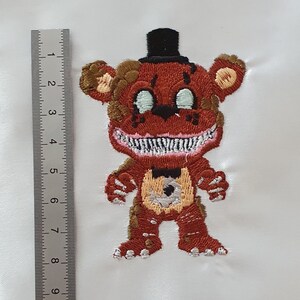 Può includere: Toppa ricamata di un orso rosso con un cappello a cilindro nero, occhi bianchi e un ampio sorriso. L'orso è in piedi con le braccia distese.