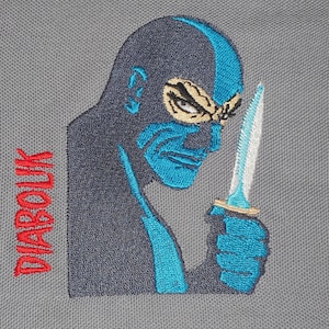Può includere: Toppa ricamata con un ninja blu e nero che tiene un coltello argentato. Il testo "DIABOLIK" è ricamato in rosso sul lato sinistro della toppa.