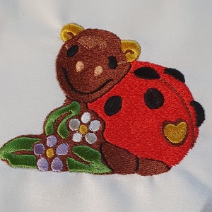 Può includere: Disegno ricamato di una coccinella marrone con macchie nere e un guscio rosso, seduta su una foglia verde con fiori viola e bianchi.