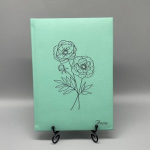 May include: A mint green journal with a black line drawing of two flowers and the name "Anna" printed on the front.