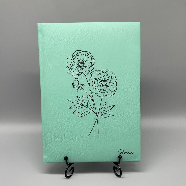 May include: A mint green journal with a black line drawing of two flowers and the name "Anna" printed on the front.