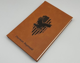 May include: A brown leather journal with an eagle and American flag design engraved on the front cover. The name "Darman Reginald" is also engraved on the cover.