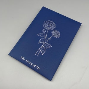 May include: A navy blue journal with a white floral design and the text "The Story of Us" on the front cover.