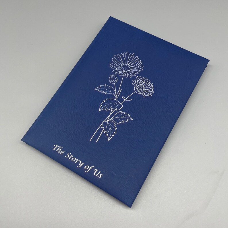 May include: A navy blue journal with a white floral design and the text "The Story of Us" on the front cover.