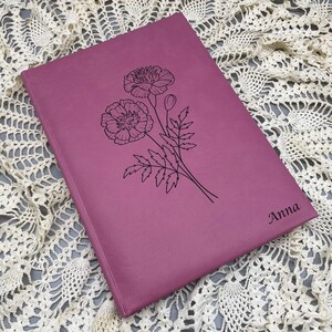 May include: A pink leather-bound journal with a floral design and the name "Anna" engraved on the cover.