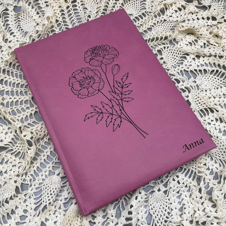 May include: A pink leather-bound journal with a floral design and the name "Anna" engraved on the cover.