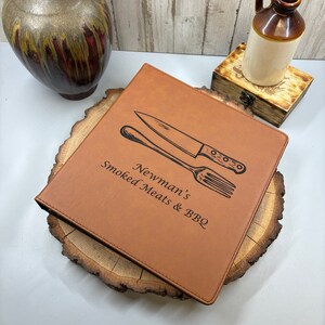 May include: A brown leather recipe binder with a black engraved design of a knife and fork. The text "Newman's Smoked Meats & BBQ" is engraved on the front.