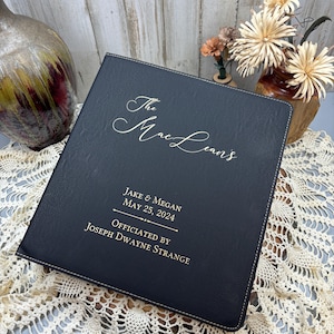 May include: Black leather wedding guest book with gold lettering. The text reads "The Mae Leans, Jake & Megan, May 25, 2024, Officiated by Joseph Dwayne Strange."