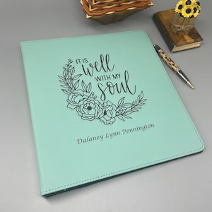 May include: A light teal binder with the text "IT IS well WITH MY soul" in a floral wreath design. The name "Dalaney Lynn Pennington" is printed below the text. A pen and small decorative items are in the background.