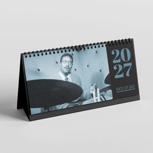 Könnte beinhalten: Ein schwarzer Tischkalender mit einem Schwarz-Weiß-Bild eines Jazzmusikers, der Schlagzeug spielt. Der Kalender zeigt das Jahr 2027 und den Text "FACES OF JAZZ". Der Kalender ist spiralgebunden und steht aufrecht.