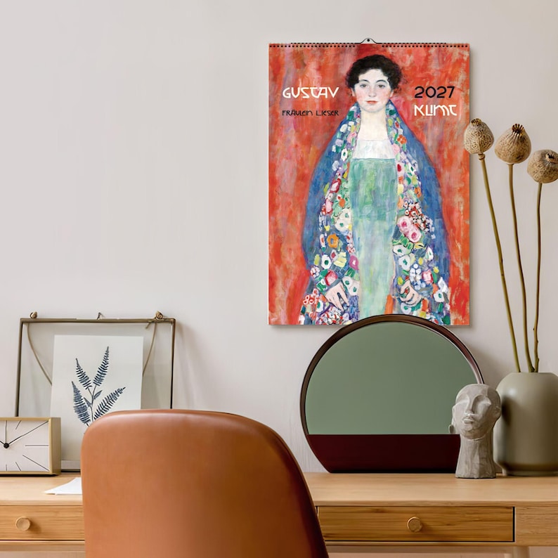 A 2027 wall calendar featuring Klimt's "Fräulein Lieser" hangs above a wooden desk with a brown chair, a clock, and a round mirror.