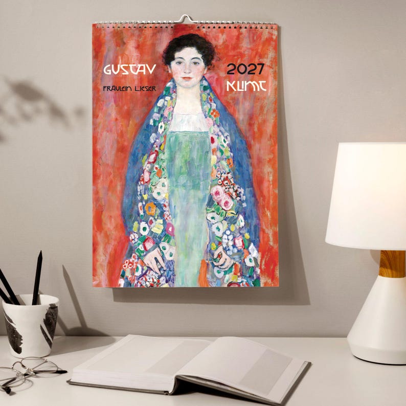 A 2027 wall calendar featuring Klimt's "Fräulein Lieser" hangs above an open book on a wooden desk with a white lamp and pens.