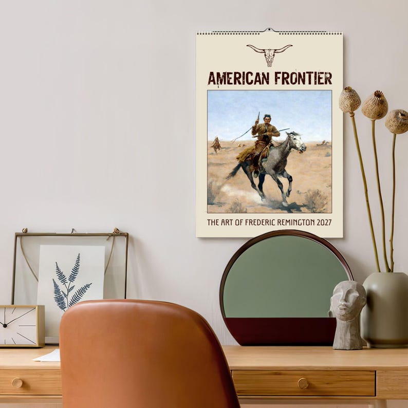 A wall calendar with a sepia-toned image of a cowboy on a horse in a desert landscape, titled "AMERICAN FRONTIER" and "THE ART OF FREDERIC REMINGTON 2027", hangs on a wall above a wooden desk with a mirror and decorative items.