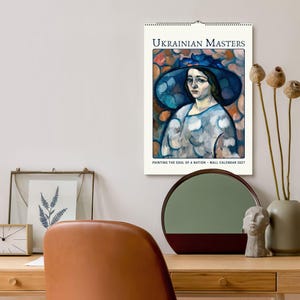 Może przedstawiać: Kalendarz ścienny zatytułowany UKRAINIAN MASTERS PAINTING THE SOUL OF A NATION - WALL CALENDAR 2027. Kalendarz zawiera drukowaną reprodukcję obrazu przedstawiającego osobę w dużym niebieskim kapeluszu i jasnej bluzce z wzorami.