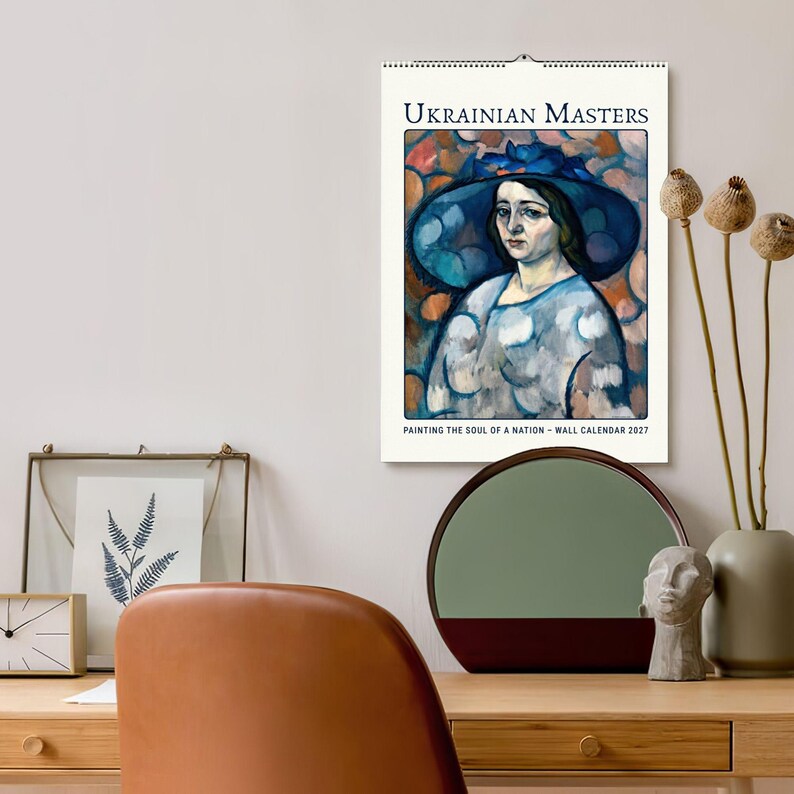 Może przedstawiać: Kalendarz ścienny zatytułowany UKRAINIAN MASTERS PAINTING THE SOUL OF A NATION - WALL CALENDAR 2027. Kalendarz zawiera drukowaną reprodukcję obrazu przedstawiającego osobę w dużym niebieskim kapeluszu i jasnej bluzce z wzorami.