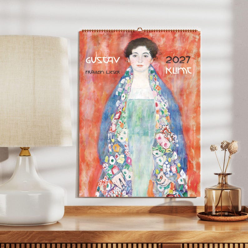 A 2027 wall calendar featuring Gustav Klimt's "Fräulein Lieser" hangs on a light gray wall above a wooden desk with a white lamp and a brown vase.