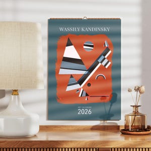 Op de afbeelding: Een wandkalender met een abstract kunstontwerp van Wassily Kandinsky. De kalender heeft een teal achtergrond met een oranje vorm in het midden. De kunst bevat geometrische vormen in wit, zwart en grijs. De tekst "WASSILY KANDINSKY" en "ART CALENDAR 2026" zijn gedrukt.