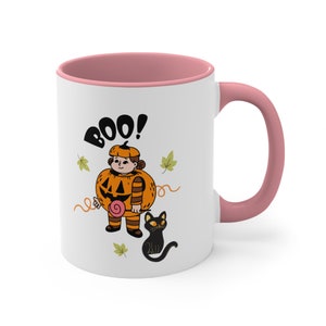 May include: A white ceramic mug with a pink rim and handle. The mug features a graphic of a child in a pumpkin costume holding a pink lollipop, with the text BOO! above them. A black cat sits nearby, surrounded by small green autumn leaves.